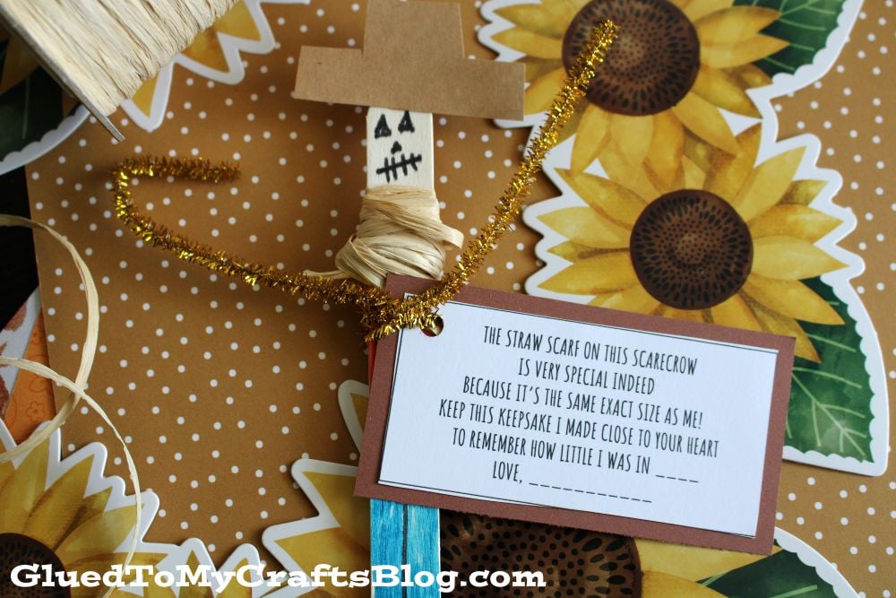 A handmade scarecrow craft with a note attached, decorated with sunflower patterns. The note explains the significance of the scarecrows scarf and has a space for a childs name.