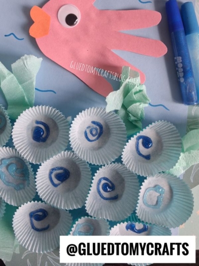 A children’s ocean craft with a pink handprint fish, waves, blue glitter “pearls” in mini cups as oysters, green tissue as seaweed, and the text @GLUEDTOMYCRAFTS at the bottom.