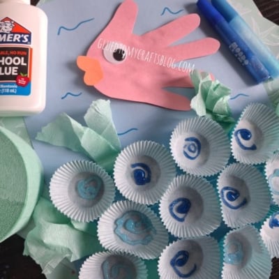 Children’s ocean craft with a paper hand fish, blue markers, cupcake liners as shells, tissue paper, school glue, and blue cupcake wrappers on a blue background. @GLUEDTOMYCRAFTS is at the bottom.