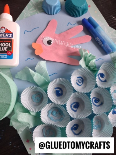 Children’s ocean craft with a paper hand fish, blue markers, cupcake liners as shells, tissue paper, school glue, and blue cupcake wrappers on a blue background. @GLUEDTOMYCRAFTS is at the bottom.