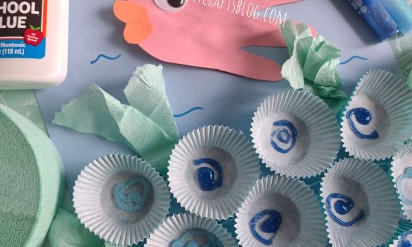 Children’s ocean craft with a paper hand fish, blue markers, cupcake liners as shells, tissue paper, school glue, and blue cupcake wrappers on a blue background. @GLUEDTOMYCRAFTS is at the bottom.