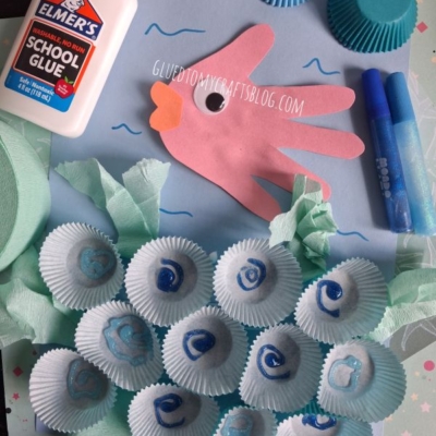 A pink handprint paper fish, glue, markers, blue cupcake liners, and a paper ocean scene with tissue paper seaweed and cupcake liner seashells, displayed on a crafting surface. @GLUEDTOMYCRAFTS watermark is visible.