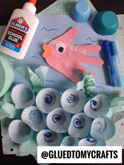A pink handprint paper fish, glue, markers, blue cupcake liners, and a paper ocean scene with tissue paper seaweed and cupcake liner seashells, displayed on a crafting surface. @GLUEDTOMYCRAFTS watermark is visible.
