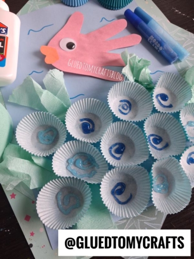 A sea-themed kids craft scene with a pink handprint fish, blue cupcake liners decorated with waves, tissue paper, glue, and markers. The text @GLUEDTOMYCRAFTS is displayed at the bottom.