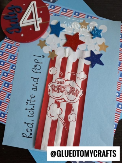 A handmade July 4th craft features a red and white popcorn bag with coding label crafts touches—star-shaped cutouts in red, white, and blue spilling out—on a blue background with patriotic paper and “Red, white and pop!” written beside it.