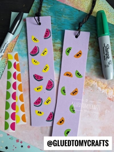 Two lavender bookmarks with colorful watermelon and kiwi stickers are on a pastel background, alongside strips of dot stickers, black and green Sharpie markers, and a coding label crafts vibe. The Instagram handle @GLUEDTOMYCRAFTS is visible.
