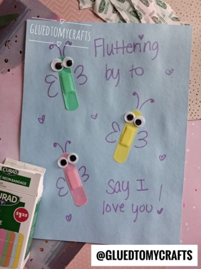 Blue paper with three butterfly crafts made from colorful Band-Aids, googly eyes, and marker. Handwritten note reads, Fluttering by to say I love you! This charming Band-Aid Butterfly Craft features SURGICAL bandages and @GLUEDTOMYCRAFTS visible. Hearts drawn on paper.