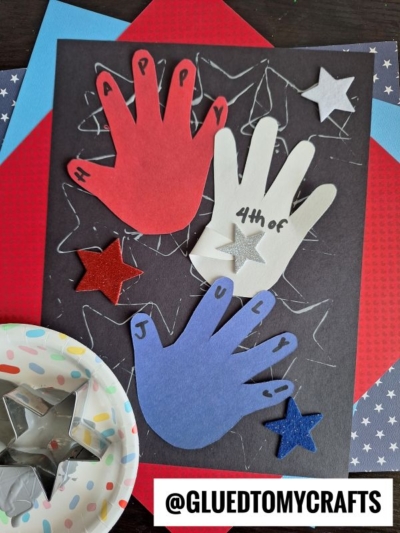 A patriotic craft featuring red, white, and blue handprint cutouts on black paper, decorated with stars for unique 4th of July keepsakes. A small bowl with star-shaped tools and @GLUEDTOMYCRAFTS is visible.