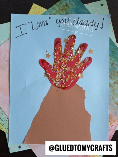 A handmade HANDPRINT FATHER'S DAY card with a brown paper volcano and red handprint lava, decorated with glitter and star confetti. "I Lava you Daddy!" is written on blue paper. @GLUEDTOMYCRAFTS is at the bottom.