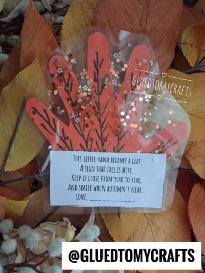 A handprint-shaped orange paper leaf with glitter sits on autumn leaves. A note reads: This little hand became a leaf, a sweet handprint leaf poem and sign that fall is here… Love, ____. The username @GLUEDTOMYCRAFTS appears at the bottom.