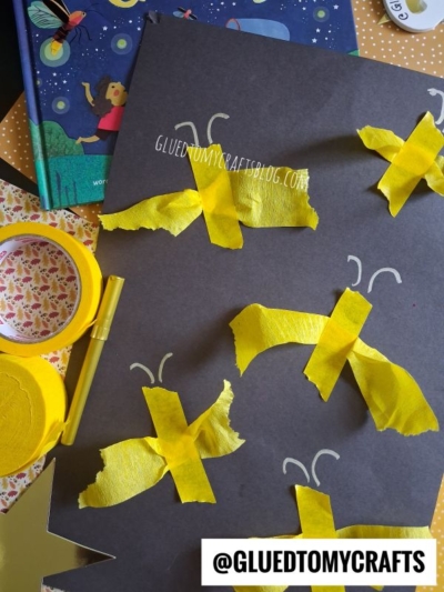 A black paper with yellow tape fireflies, white marker wings, and craft supplies like yellow tape, a book, and a yellow pen around it. @GLUEDTOMYCRAFTS appears in the corner.
