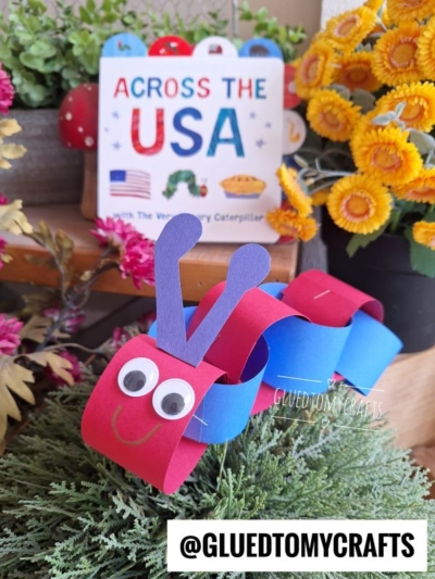 A red and blue paper caterpillar craft—perfect for 4th of July crafts—with googly eyes and purple antennae sits on green foliage. Behind it is a book titled Across the USA, bright yellow artificial flowers, and @GLUEDTOMYCRAFTS.