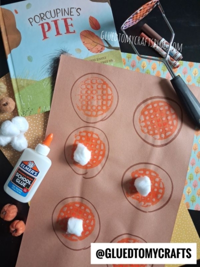 Children’s craft setup: a Porcupine’s Pie book, dotted paint roller, glue, paper with pie drawings, cotton balls, orange pom-poms, and patterned paper on a table—perfect for a fun potato masher stamped craft activity. @GLUEDTOMYCRAFTS is labeled at the bottom.