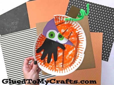 A hand holds a Halloween craft made from a paper plate pumpkin with cut-out eyes, a handprint, and a green pipe cleaner stem. The background has patterned scrapbook paper.