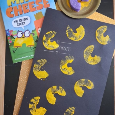 Childrens book Mac & Cheese: The Origin Story, a donut with yellow paint, and black paper with pool noodle stamped mac and cheese shapes; @GLUEDTOMYCRAFTS is written at the bottom.