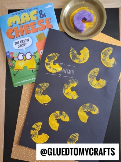 Childrens book Mac & Cheese: The Origin Story, a donut with yellow paint, and black paper with pool noodle stamped mac and cheese shapes; @GLUEDTOMYCRAFTS is written at the bottom.