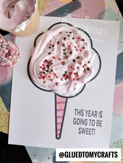A paper craft of a pink ice cream cone with sprinkles sits on a card that reads This year is going to be sweet! Assorted craft materials—including puffy paint and a cup of pink paste that looks just like cotton candy—are nearby. @GLUETOMYCRAFTS is written twice.