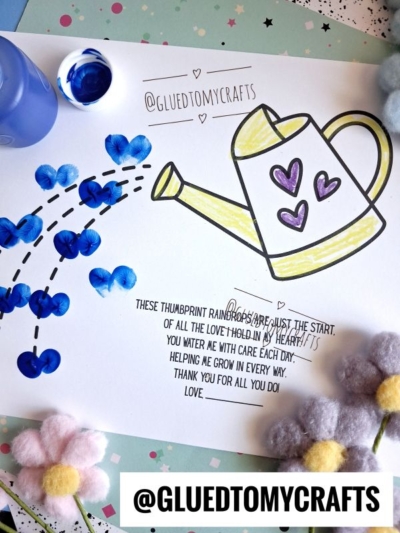 A printable craft sheet shows a thumbprint watering can craft outlined in black, decorated with purple hearts. Blue paint thumbprints form a “rainbow” of water droplets, while felt flowers and an open paint pot are nearby.