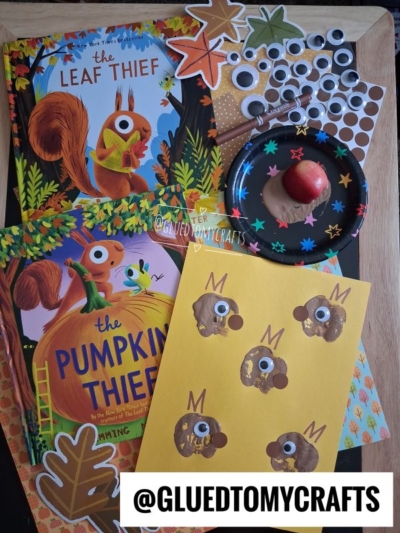 Two childrens books, autumn-themed stickers, a paper plate with brown paint and a red apple, and a yellow sheet with squirrel crafts sit on a wooden surface. A @GLUEDTOMYCRAFTS label appears at the bottom.