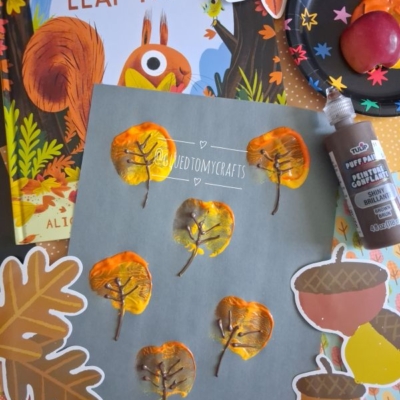 Children’s autumn leaf art project using paint and glue is shown on gray paper, surrounded by a fall-themed book, fake leaves, and a paint palette. The @gluedtomycrafts handle is at the bottom.