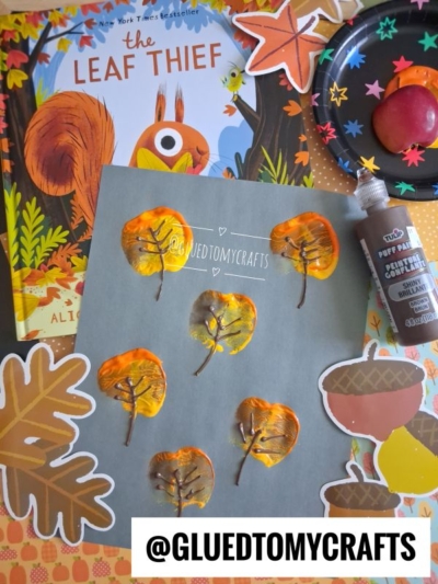 Children’s autumn leaf art project using paint and glue is shown on gray paper, surrounded by a fall-themed book, fake leaves, and a paint palette. The @gluedtomycrafts handle is at the bottom.