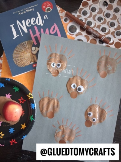 Children’s craft project featuring brown handprint hedgehogs on gray paper, googly eyes attached, the book “I Need a Hug,” a brown marker, and an apple in a black paper plate—perfect for an APPLE STAMPED CRAFT—along with the handle of a glue stick visible.
