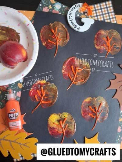 Six apple stamped leaves, created by printing with apple halves painted in rich fall colors, are displayed on black paper. A red apple, paint, and fall-themed paper leaves add to the cozy autumn vibe. The username @GLUEDTOMYCRAFTS is featured.