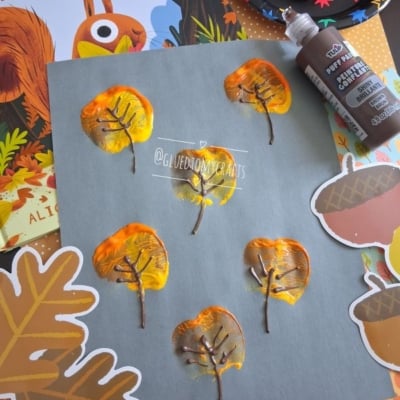 A gray paper with leaf prints made from painted apple halves, brown paint for branches, autumn-themed stickers, and a book titled The Leaf Thief in the background. @GLUEDTOMYCRAFTS watermark is visible.