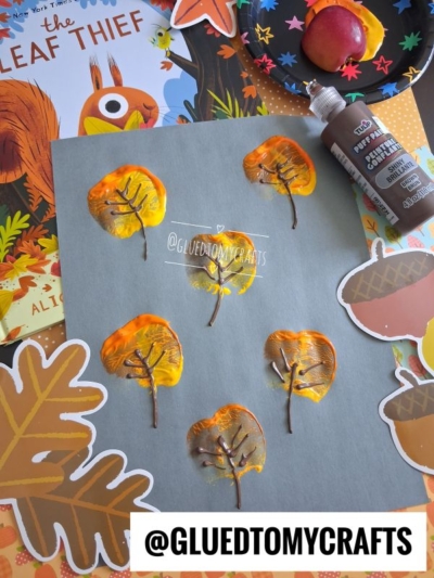A gray paper with leaf prints made from painted apple halves, brown paint for branches, autumn-themed stickers, and a book titled The Leaf Thief in the background. @GLUEDTOMYCRAFTS watermark is visible.