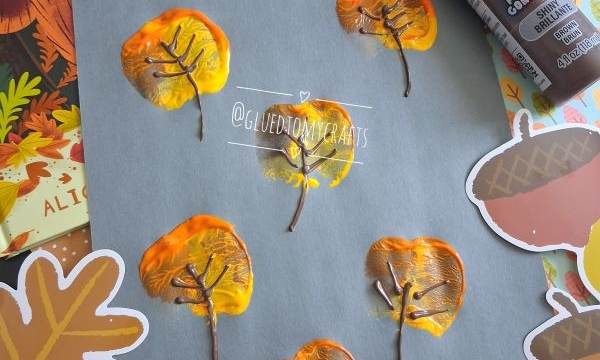 A gray paper with leaf prints made from painted apple halves, brown paint for branches, autumn-themed stickers, and a book titled The Leaf Thief in the background. @GLUEDTOMYCRAFTS watermark is visible.