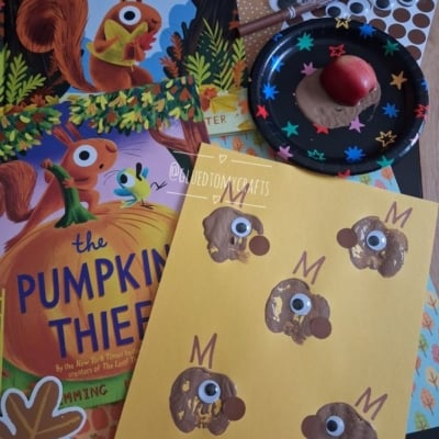 Children’s book The Leaf Thief, The Pumpkin Thief, squirrel crafts with googly eyes, brown paint noses, and a small apple on a black plate. Craft materials are spread on a table.