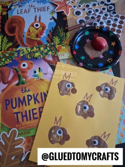 Children’s book The Leaf Thief, The Pumpkin Thief, squirrel crafts with googly eyes, brown paint noses, and a small apple on a black plate. Craft materials are spread on a table.