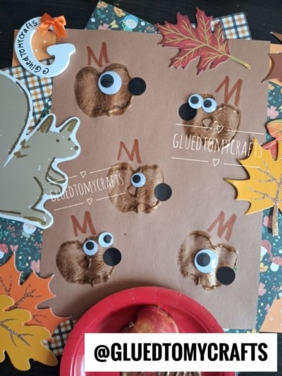 A brown sheet with apple stamp prints—some transformed into moose faces using googly eyes and paper antlers, others forming the body of an apple stamped squirrel—creates an autumn woodland scene. Surrounded by colorful leaves, a playful squirrel cutout, and a red plate with an apple. Text reads @GLUETOMYCRAFTS.