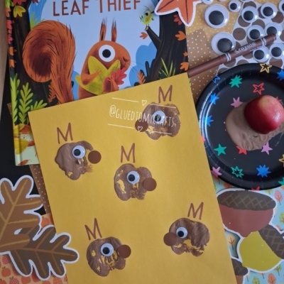 A childrens craft setup with The Leaf Thief book, brown thumbprint animals on yellow paper, googly eyes, paint, leaves, and apple, all displayed on a colorful autumn-themed background. @GLUEDTOMYCRAFTS.