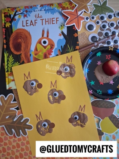 A childrens craft setup with The Leaf Thief book, brown thumbprint animals on yellow paper, googly eyes, paint, leaves, and apple, all displayed on a colorful autumn-themed background. @GLUEDTOMYCRAFTS.