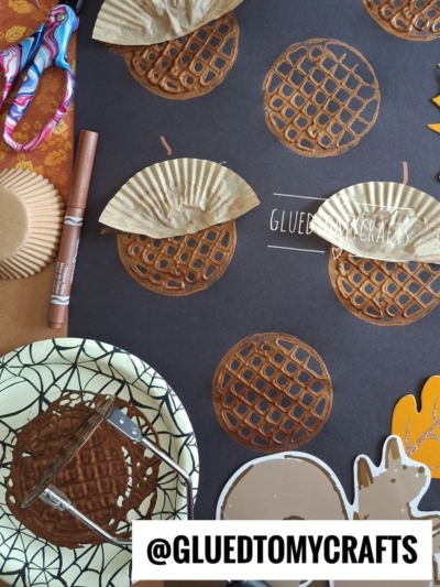 Craft project with brown cupcake liners and drawn circles on black paper, resembling waffles. Craft supplies, scissors, and a marker are nearby—perfect for pairing with a fun potato masher stamped acorn activity. Text @GLUEDTOMYCRAFTS appears at the bottom.