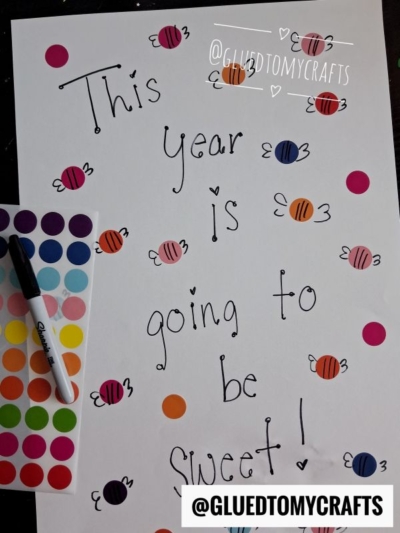 A poster reads This year is going to be sweet! with colorful candy, heart doodles, and a coding label crafts vibe. A sheet of round stickers and a black marker are nearby. The handle @GLUEDTOMYCRAFTS is written twice on the image.