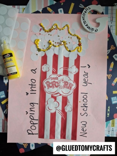 A handmade popcorn-themed back-to-school craft featuring a red and white popcorn box, yellow paint as butter, cotton popcorn, and a "Popping into a New School Year!" coding label on pink paper. @GLUETOMYCRAFTS is tagged.