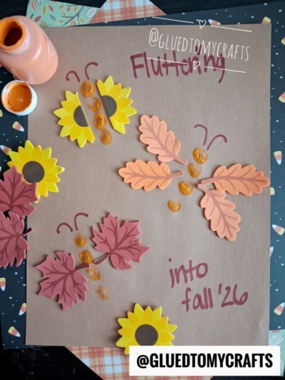 A fall-themed craft with yellow flower and orange leaf cutouts, paint fingerprints forming butterflies, and “Fluttering into fall 26” written on brown paper—perfect for FALL CRAFTS FOR KIDS. Paint and leaves decorate the edges.