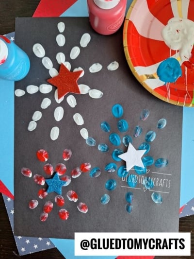 Black paper displays red, white, and blue fireworks made from painted fingerprints—a festive idea for 4th of July crafts. Each firework has a glittery star in the center. Paint bottles, a palette, and the @GLUEDTOMYCRAFTS username are also visible.