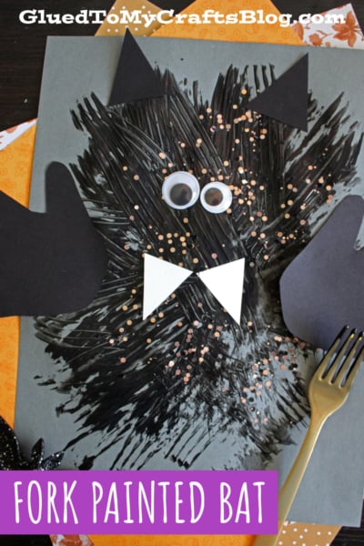 A bat made from black paint streaks using a fork, with paper ears, wings, bow tie, and googly eyes on gray paper. A gold fork and glitter are nearby. Text reads FORK PAINTED BAT.
