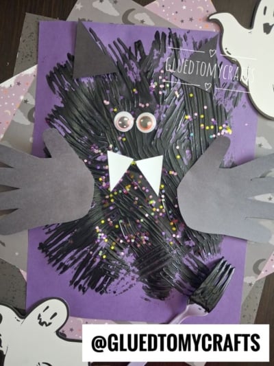 A fork painted bat craft made with black paint, googly eyes, paper wings, and fangs on purple paper. Sprinkles decorate the bat. A fork, ghosts, and the text @GLUEDTOMYCRAFTS are also visible.