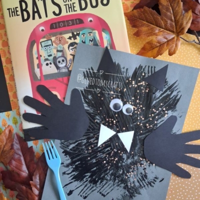 A children’s book titled “The Bats on the Bus” sits next to a fork painted bat craft made from black paint, googly eyes, paper hands, and a fork, surrounded by artificial autumn leaves. Text: @GLUEDTOMYCRAFTS.
