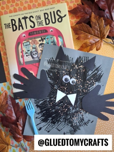 A children’s book titled “The Bats on the Bus” sits next to a fork painted bat craft made from black paint, googly eyes, paper hands, and a fork, surrounded by artificial autumn leaves. Text: @GLUEDTOMYCRAFTS.