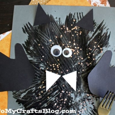 Children’s bat craft using black paint, paper cutouts for wings, ears, eyes, and bow tie, with a fork for texture. Background includes orange patterned paper.