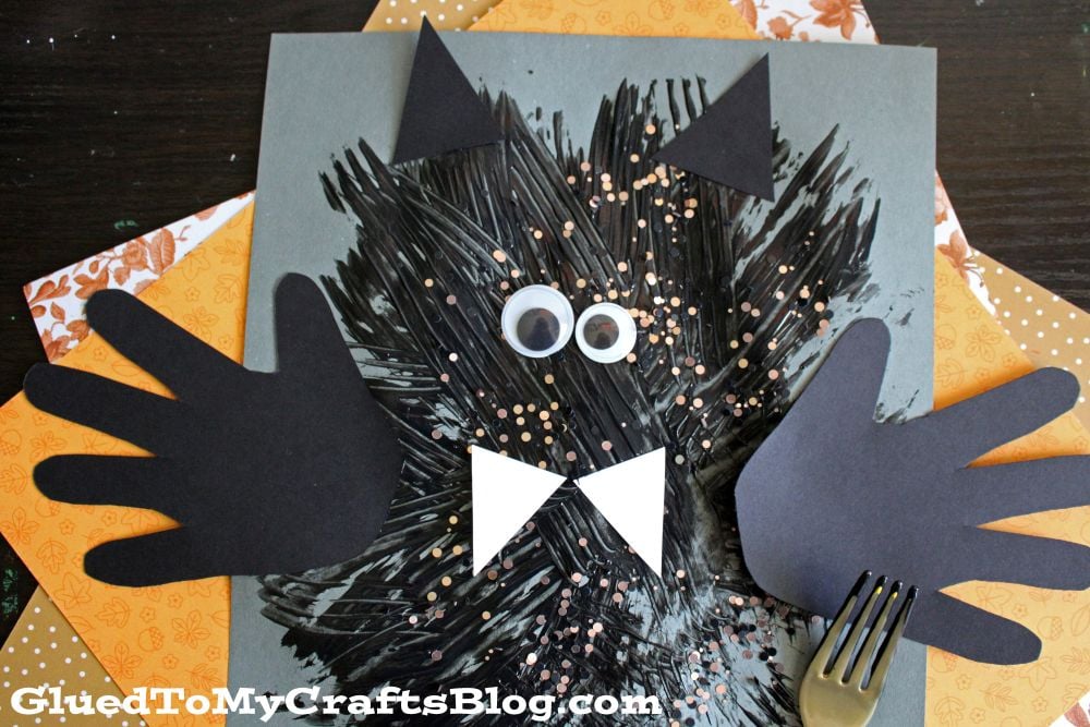 Children’s bat craft using black paint, paper cutouts for wings, ears, eyes, and bow tie, with a fork for texture. Background includes orange patterned paper.