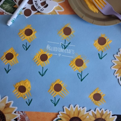 Blue paper with yellow sunflowers painted using a fork, brown centers, and green stems, surrounded by sunflower-themed napkins and a yellow plate with a purple fork and paint. @GLUEDTOMYCRAFTS is written twice.