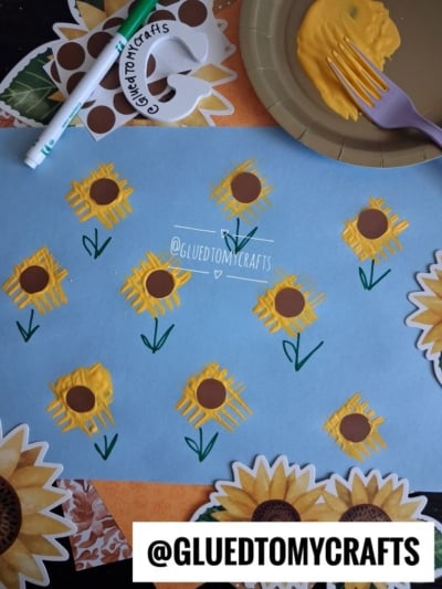 Blue paper with yellow sunflowers painted using a fork, brown centers, and green stems, surrounded by sunflower-themed napkins and a yellow plate with a purple fork and paint. @GLUEDTOMYCRAFTS is written twice.