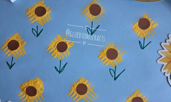 Blue paper with yellow sunflowers painted using a fork, brown centers, and green stems, surrounded by sunflower-themed napkins and a yellow plate with a purple fork and paint. @GLUEDTOMYCRAFTS is written twice.