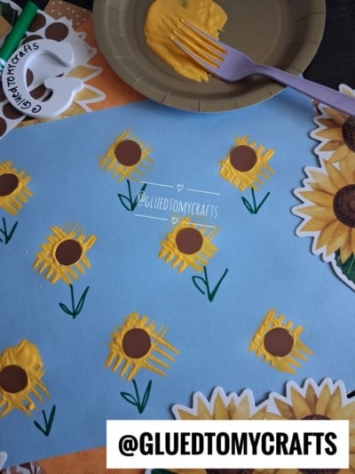 A blue paper displays sunflowers made with yellow paint and brown centers, crafted using a fork. Sunflower stickers and art materials surround the paper. @GLUEDTOMYCRAFTS watermark is visible.